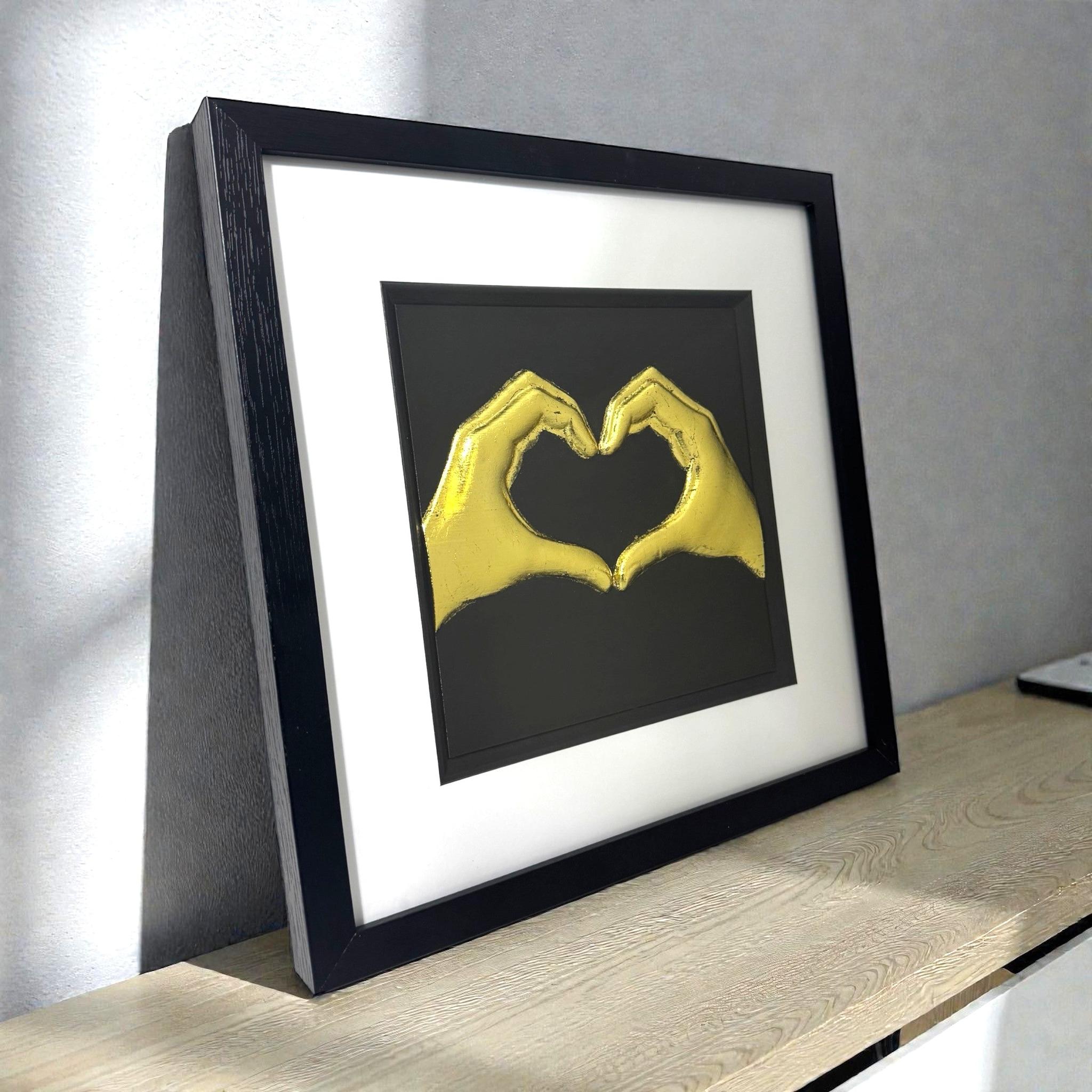 Framed love heart hands wall art in gold leaf relief on a black background, displayed on a shelf in a modern interior