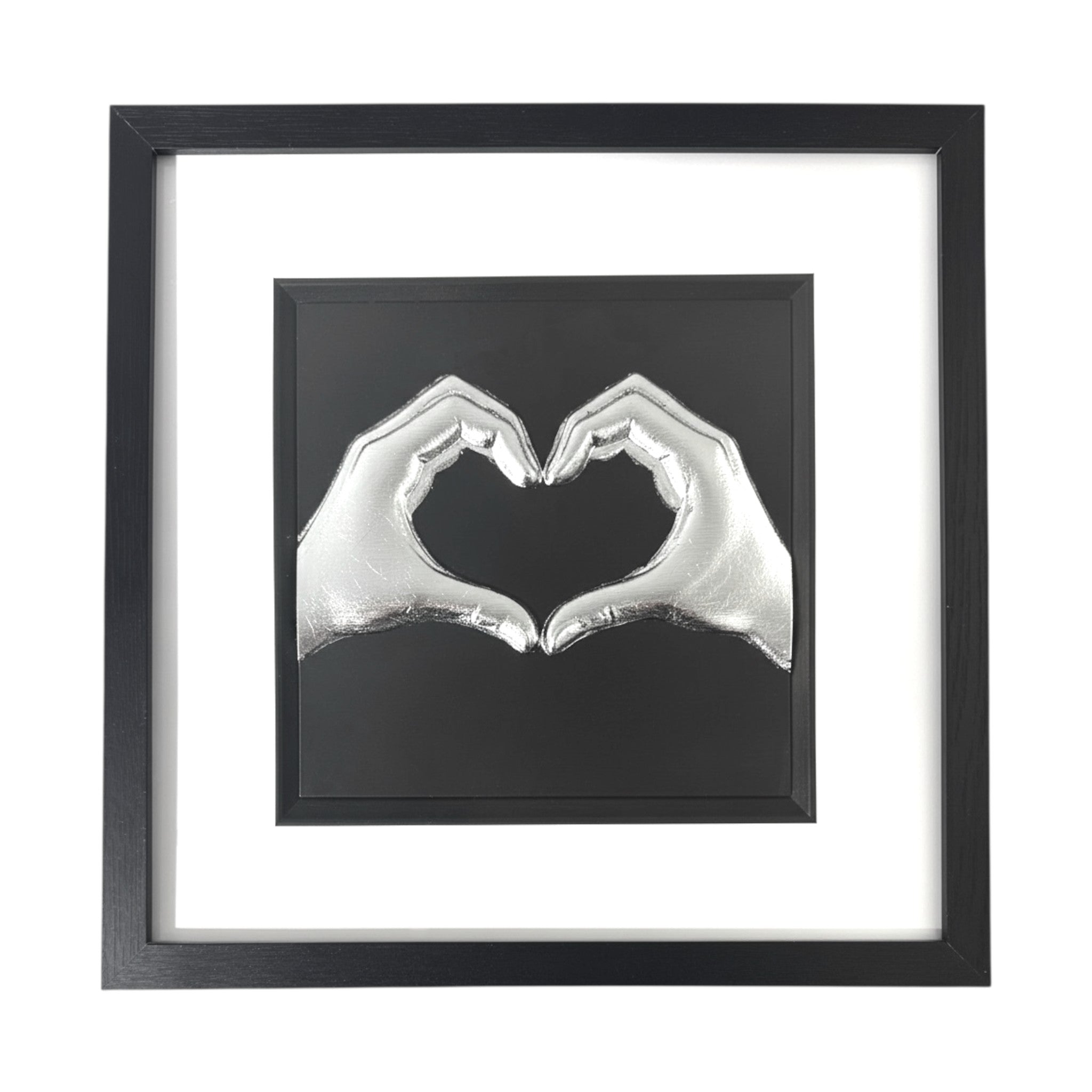 Framed love heart hands wall art in silver leaf relief, centred on a black background with white mount and black frame