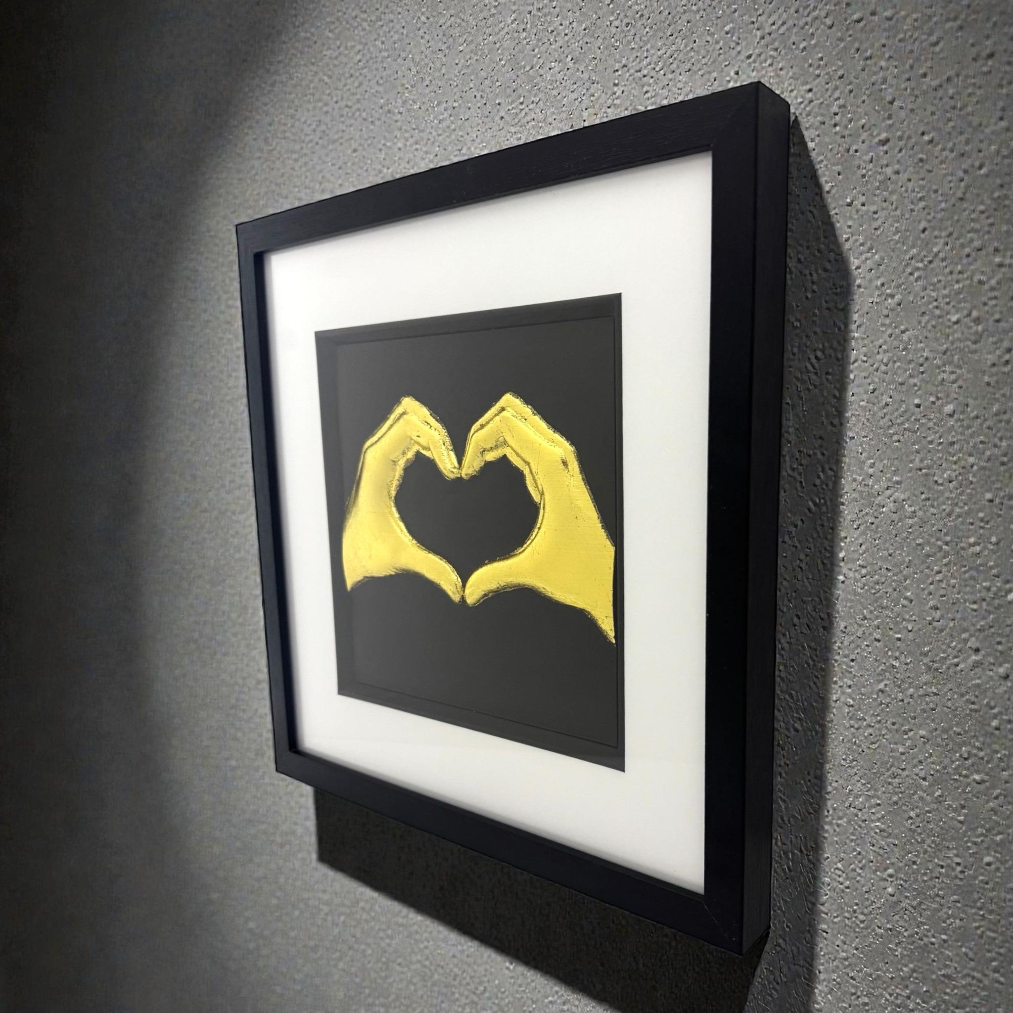 Framed love heart hands wall art in gold leaf relief mounted on a wall, shown at an angle highlighting depth and texture