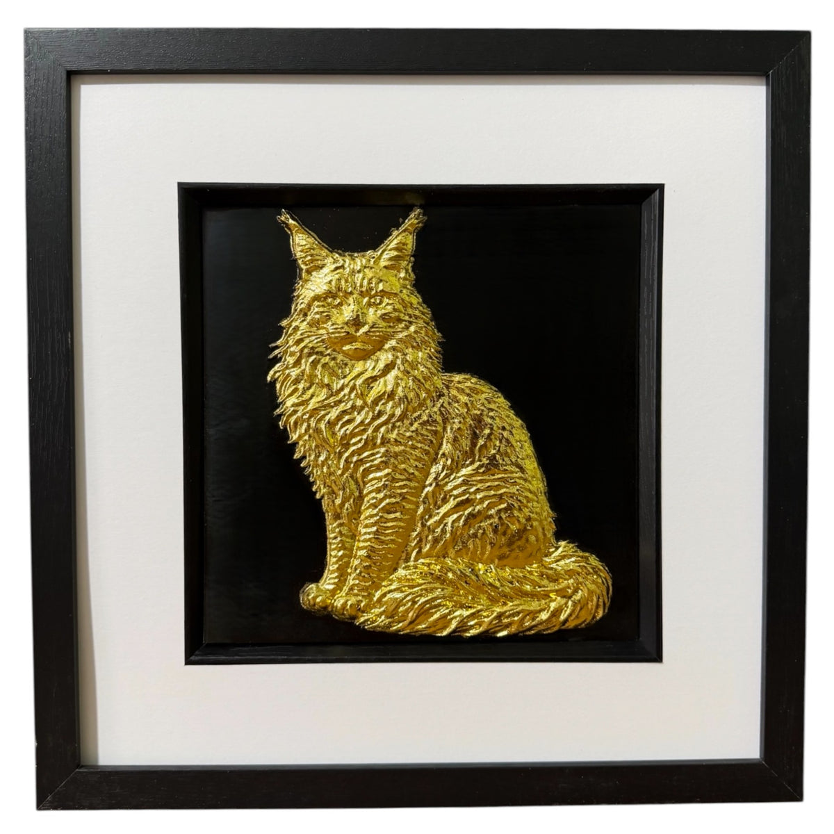 Framed Maine Coon cat wall art, gold relief of a sitting long-haired cat with tufted ears, ruff and plumed tail, on a black background in a black frame with white mount