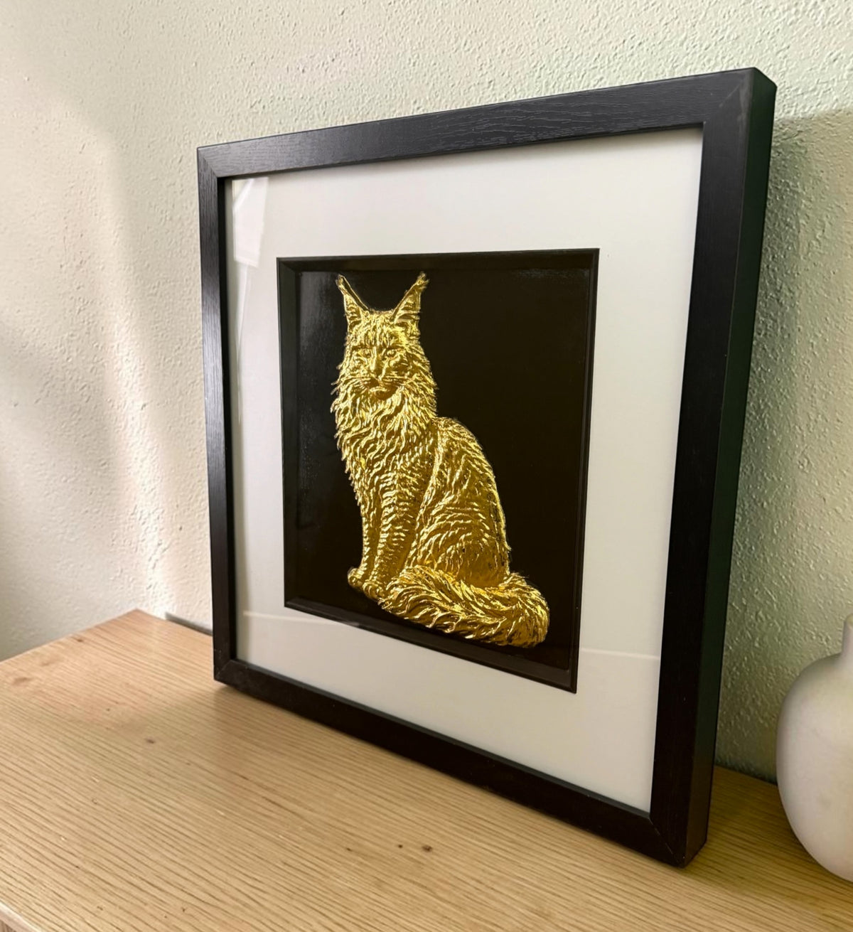 Gold Maine Coon framed wall art leaning on an oak sideboard against a pale green wall, angled to show the raised relief of the long coat through the glass