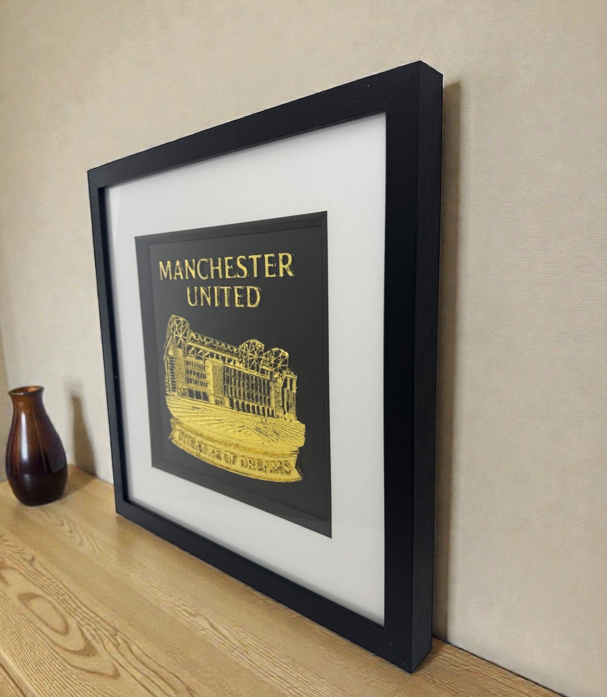 Framed Manchester United gold relief wall art on a wooden desk, black frame with white mount, stadium design with “Theatre of Dreams” text.