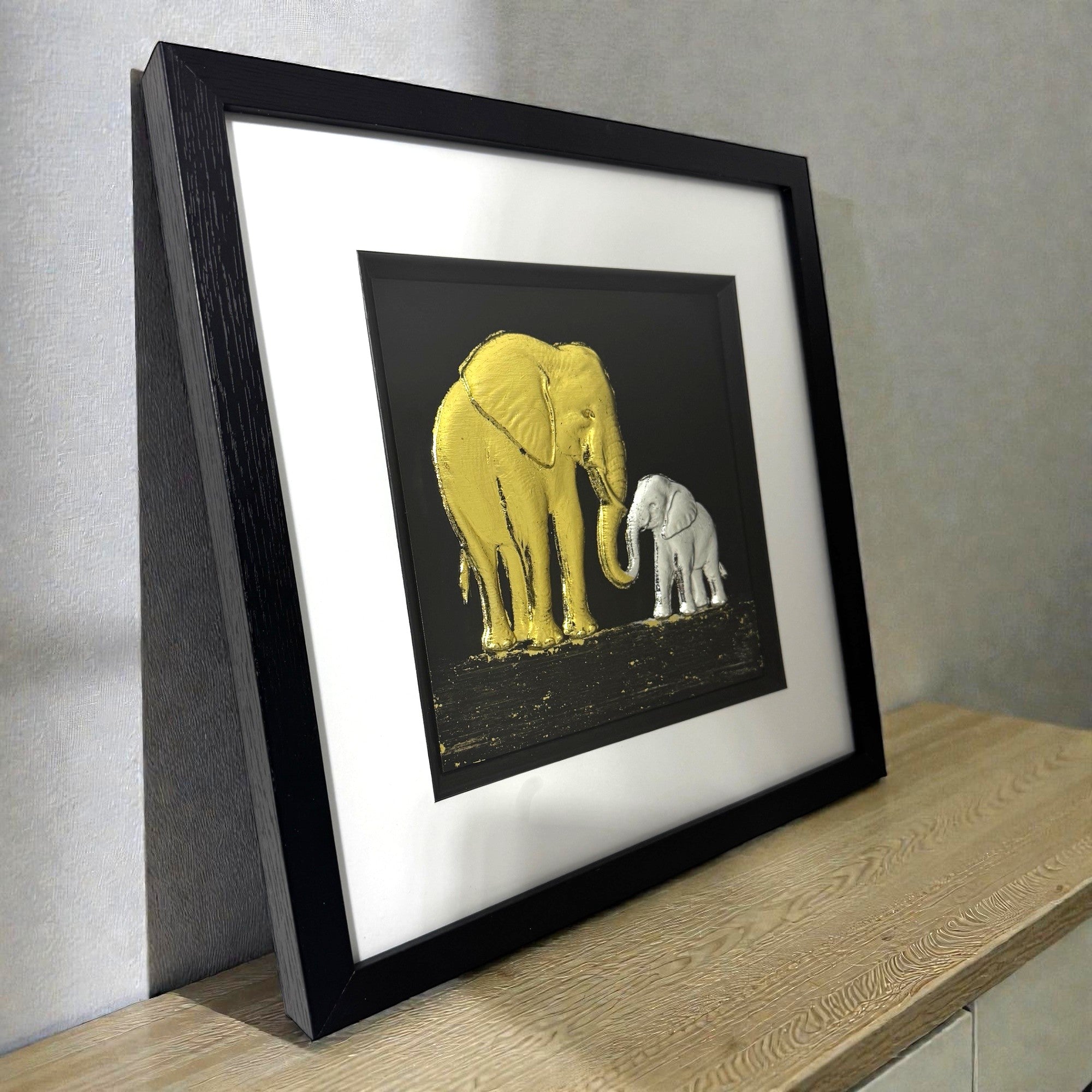 Framed elephant mother and baby wall art displayed on a shelf in a modern interior, showing gold and silver leaf relief