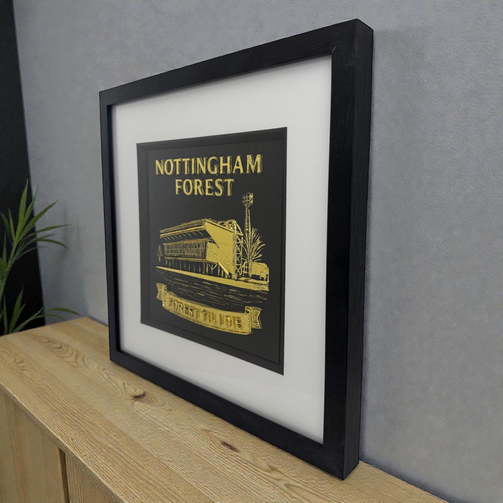 Nottingham Forest 3D wall art in gold leaf, deep black frame and white mount, lifestyle photo on wooden surface against grey wall.