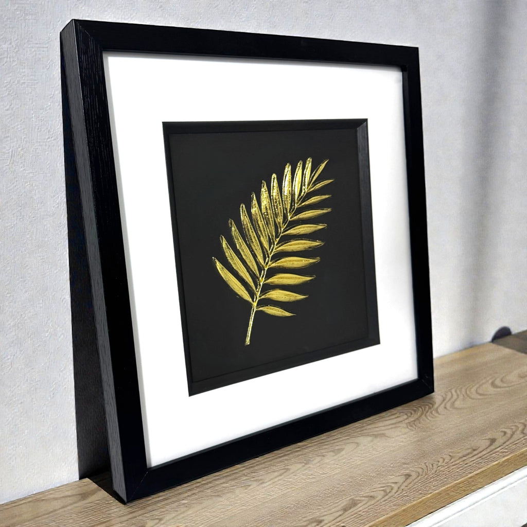 Gold palm leaf wall art shown at an angle on a wooden surface, framed with white mount and raised palm leaf relief