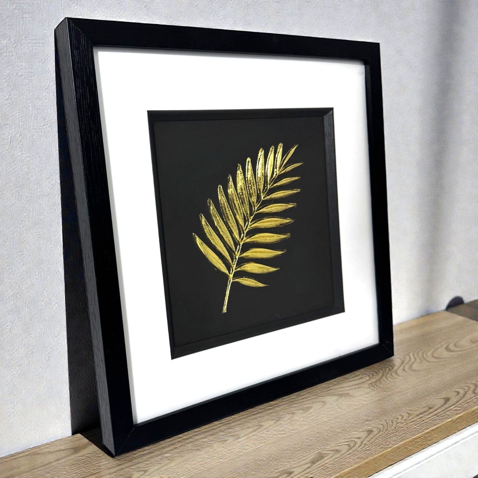 Gold palm leaf wall art shown at an angle on a wooden surface, framed with white mount and raised palm leaf relief