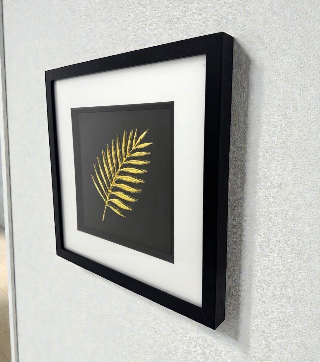 Framed gold palm leaf wall art displayed on a wall, black frame with white mount and sculpted palm frond design
