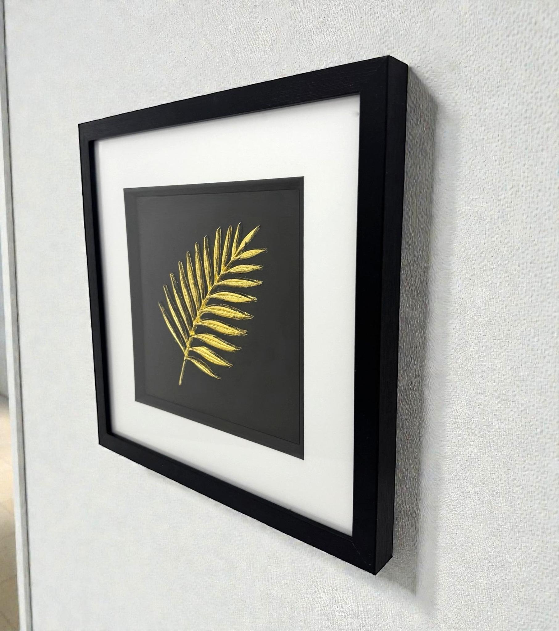 Framed gold palm leaf wall art displayed on a wall, black frame with white mount and sculpted palm frond design