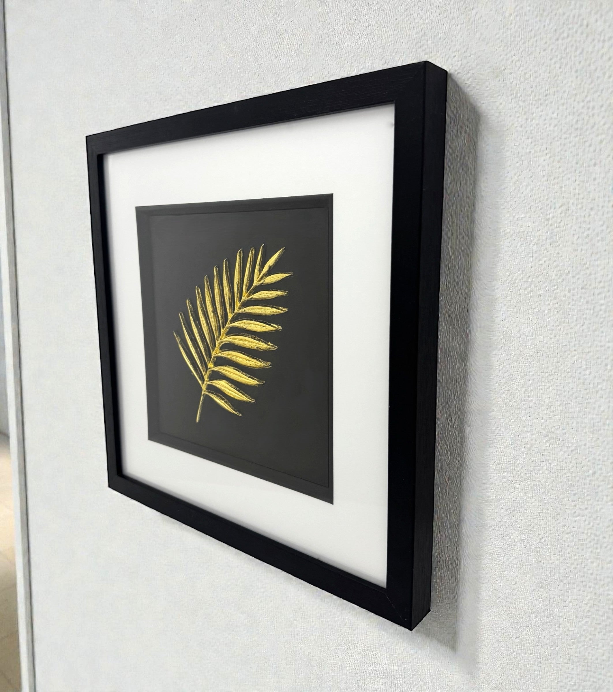 Framed gold palm leaf wall art displayed on a wall, black frame with white mount and sculpted palm frond design