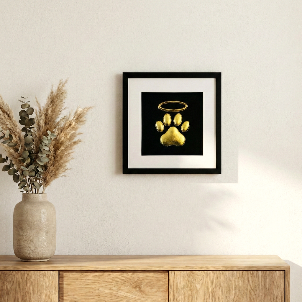 Framed dog paw print halo memorial wall art in gold leaf, styled on a neutral wall above a wooden sideboard with dried pampas and eucalyptus in a vase.