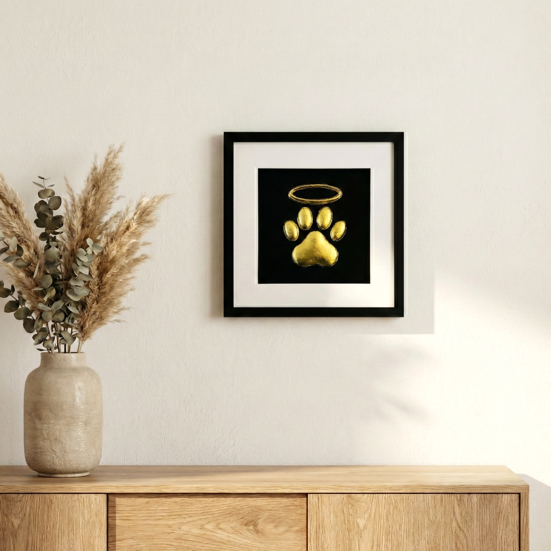 Framed dog paw print halo memorial wall art in gold leaf, styled on a neutral wall above a wooden sideboard with dried pampas and eucalyptus in a vase.