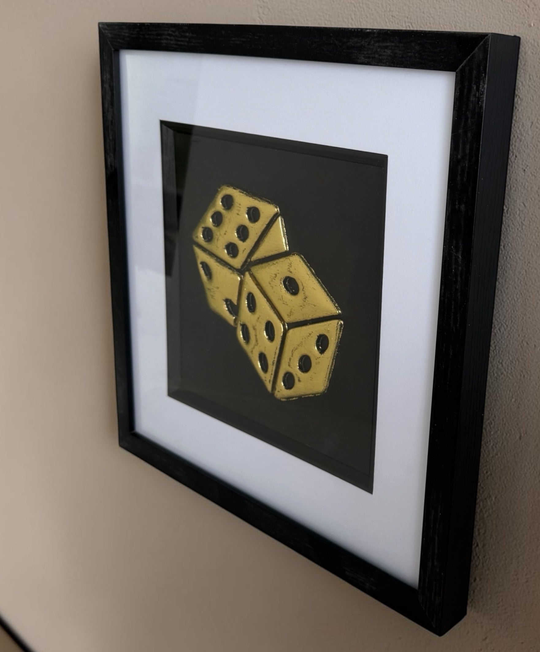 Angled close-up of framed gold leaf dice wall art with black background and white mount, shown on a wall as luxury games room decor.
