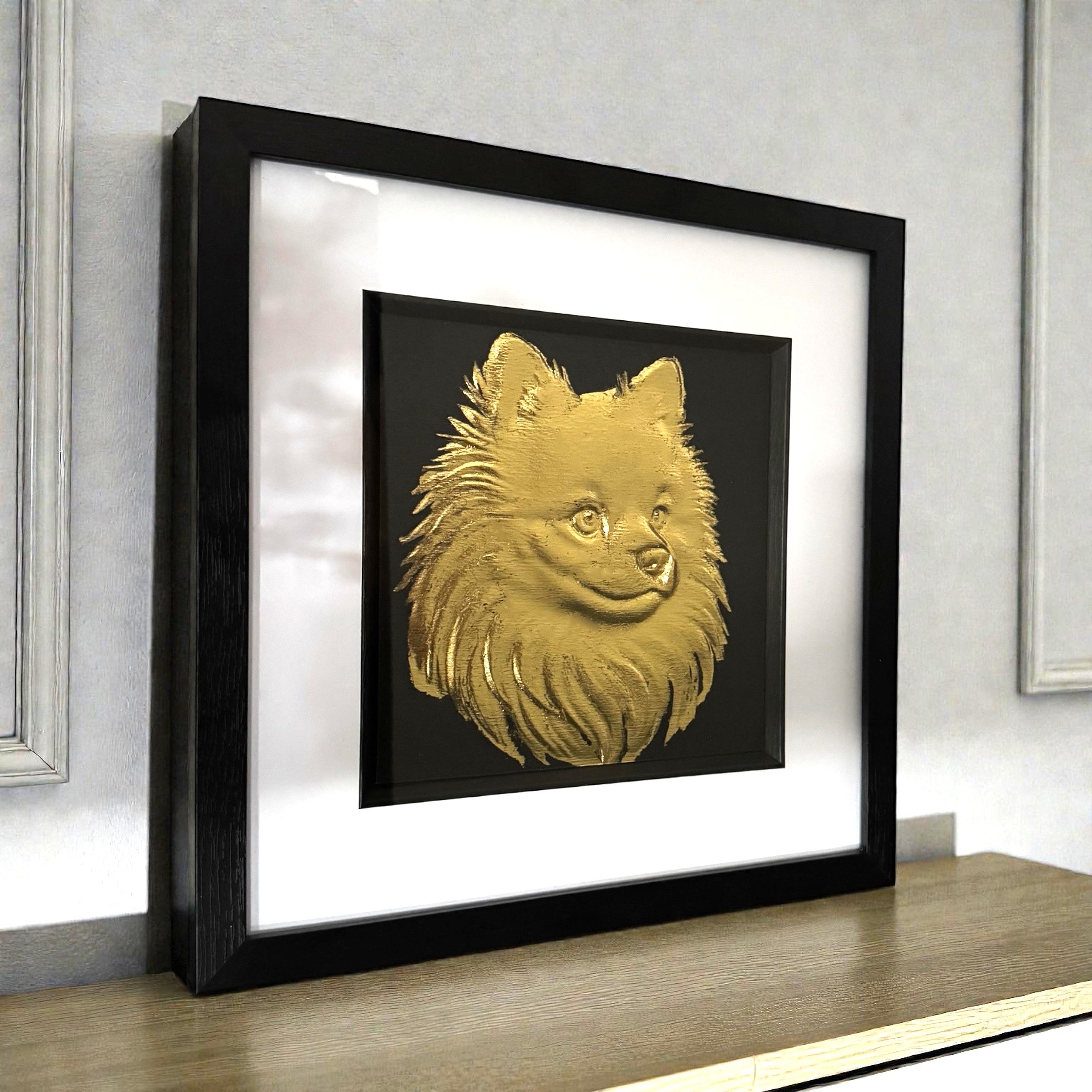 Pomeranian gold leaf framed wall art shown on a desk with black frame white mount and black background