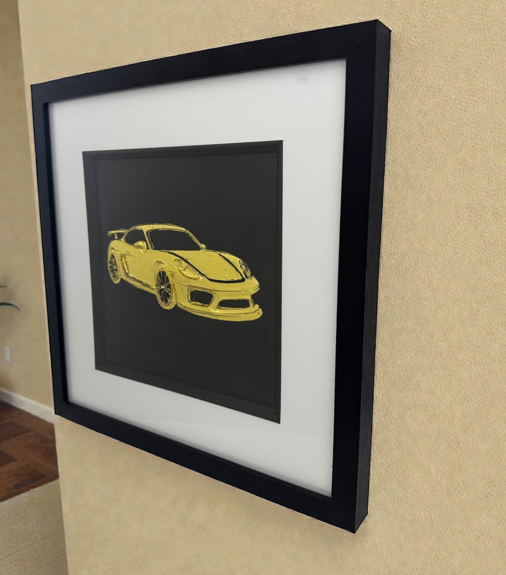 Gold leaf Porsche GT4 wall art in black frame with white mount, hung on a beige wall, 33 x 33 cm