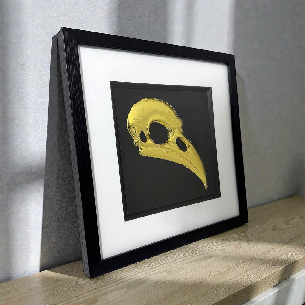Gold leaf raven skull wall art displayed on desk in black frame