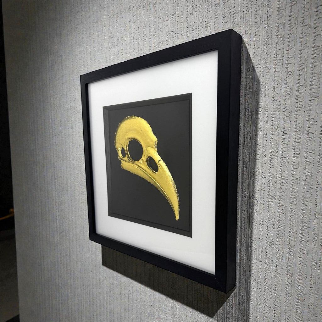 Framed raven skull wall art in gold leaf with black background and white mount