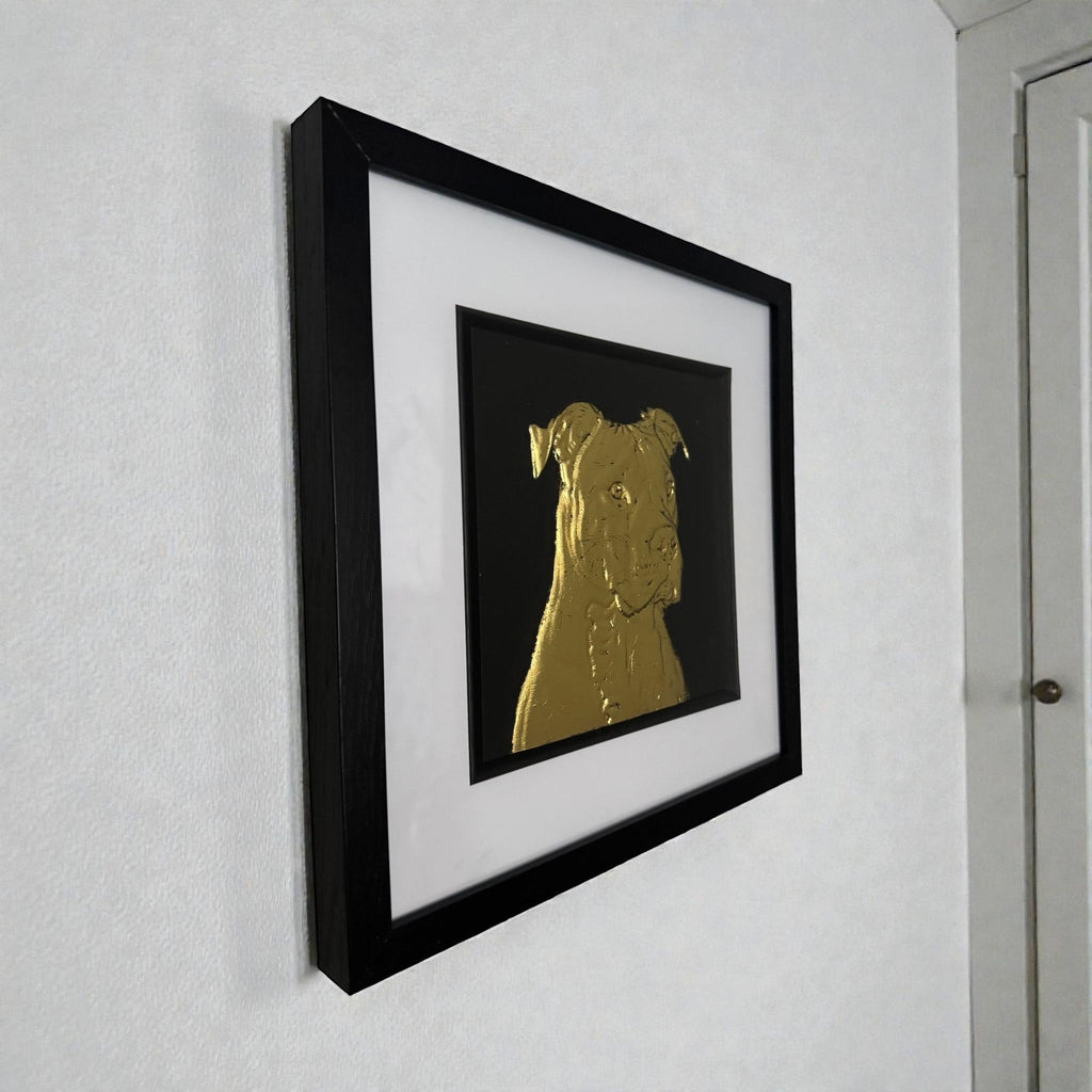 Staffordshire Bull Terrier framed wall art in gold leaf shown mounted on a wall at a slight angle with black wooden frame and white mount