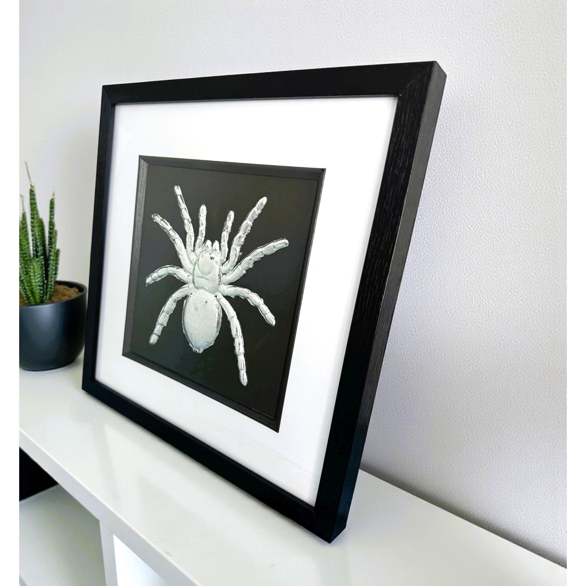 Silver tarantula wall art framed in black with white mount on a shelf