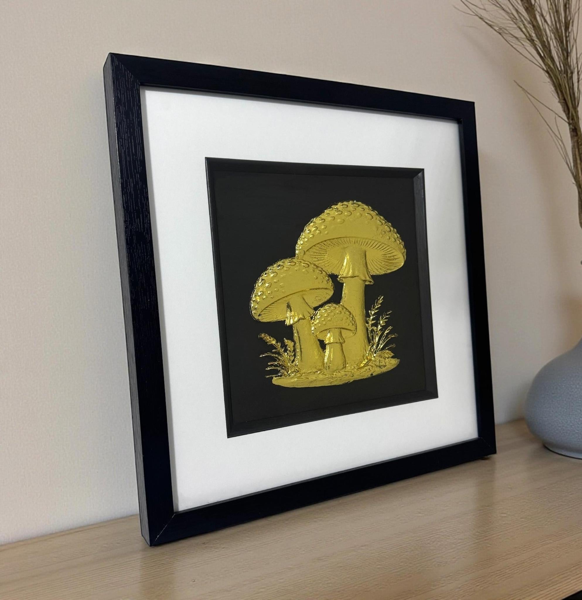 Gold leaf toadstool trio wall art in black frame with white mount, displayed on a light oak desk, 33 x 33 cm