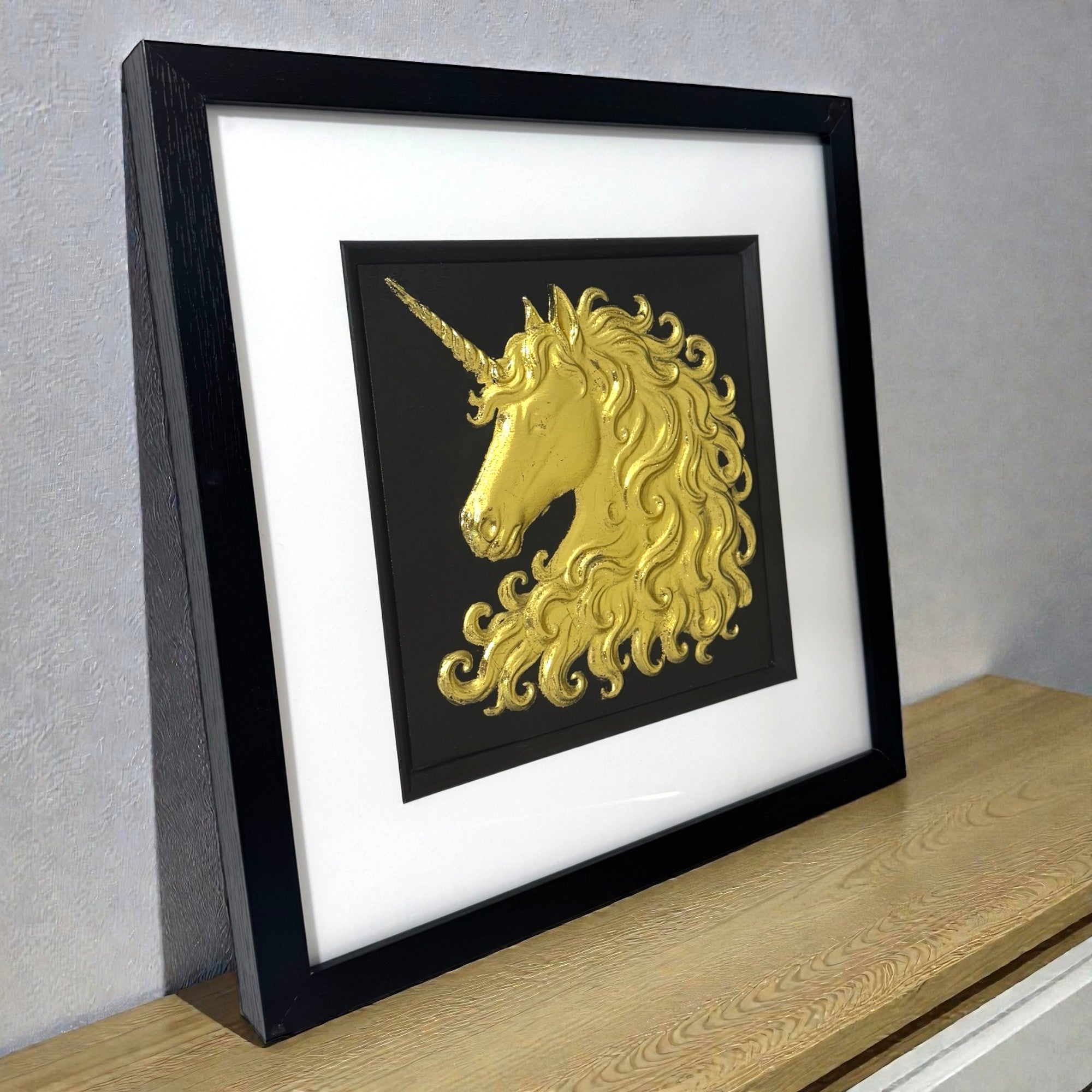 Framed unicorn wall art in gold leaf displayed on a shelf, black frame with white mount and raised relief detail