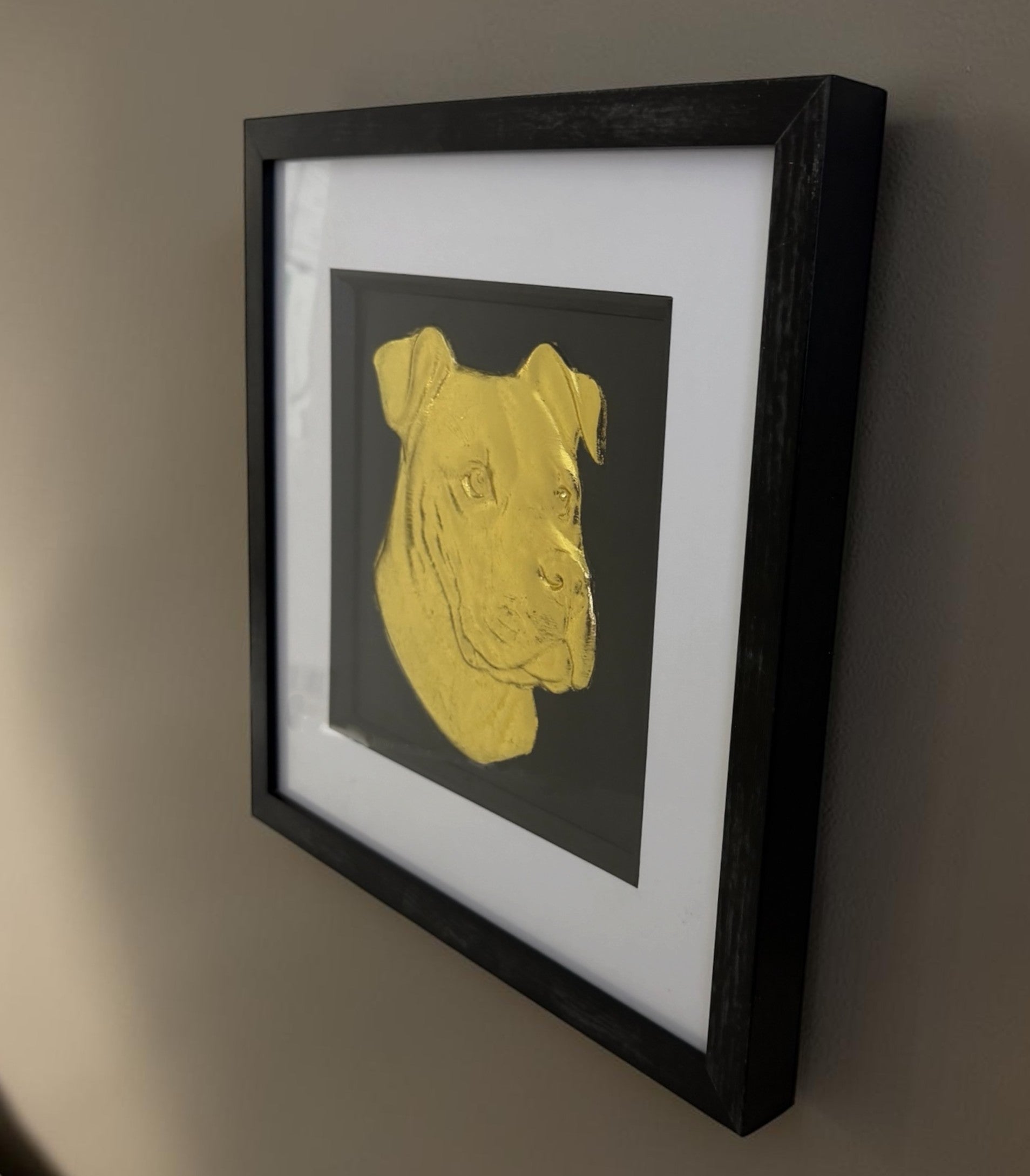 Side angle view of framed XL Bully wall art hanging on a neutral wall, featuring a gold leaf dog portrait on a black background with a black frame and white mount.