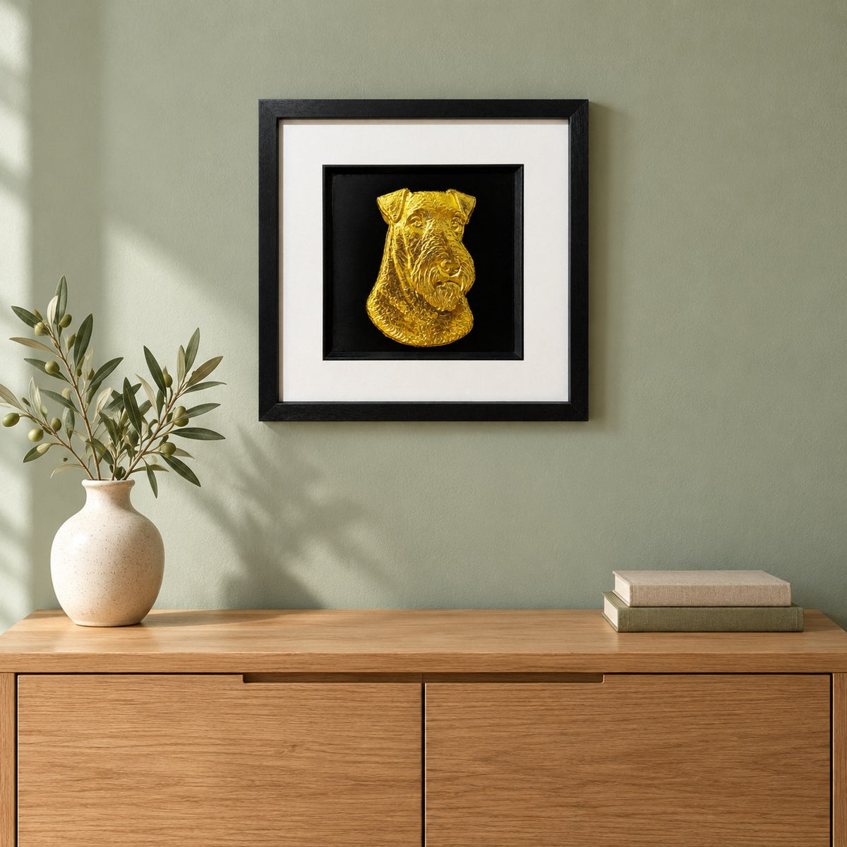 Gold Airedale Terrier 3D wall art in a black square frame hung on a sage green wall above an oak sideboard with a vase of olive branches and two linen books