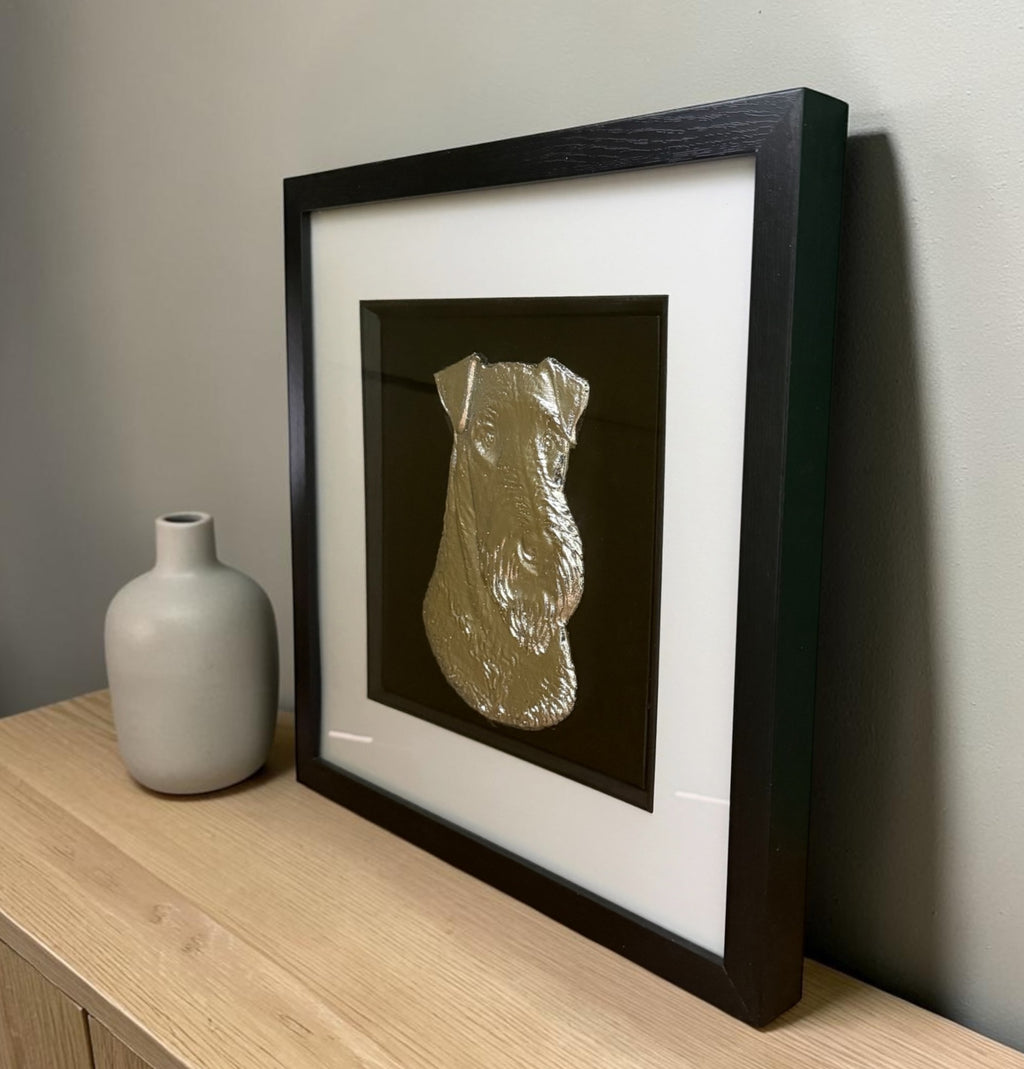 Gold Airedale Terrier framed wall art leaning on an oak sideboard beside a grey ceramic vase, angled to show the raised relief of the coat through the glass