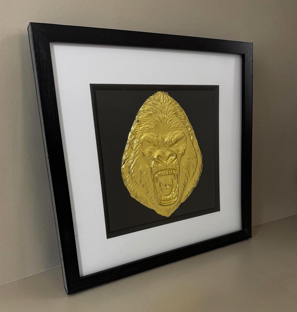 Angled lifestyle shot of the Gold finish gorilla piece leaning on a neutral surface against a neutral wall, showing frame depth and glass reflection.