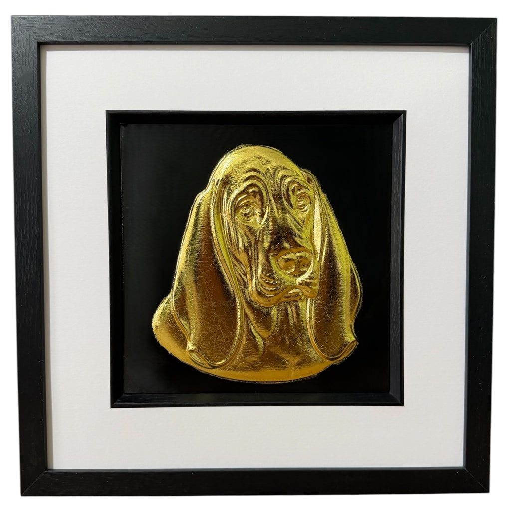 Framed Basset Hound wall art, gold relief portrait of the head and long ears on a black background, black wooden frame with white mount, on a white background