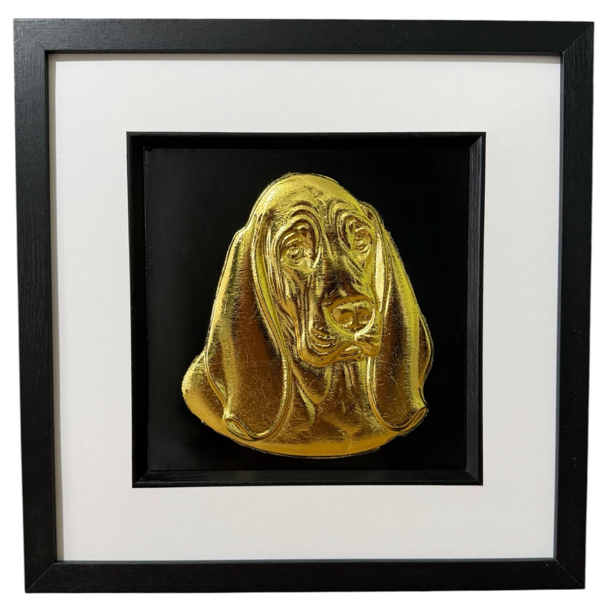 Framed Basset Hound wall art, gold relief portrait of the head and long ears on a black background, black wooden frame with white mount, on a white background