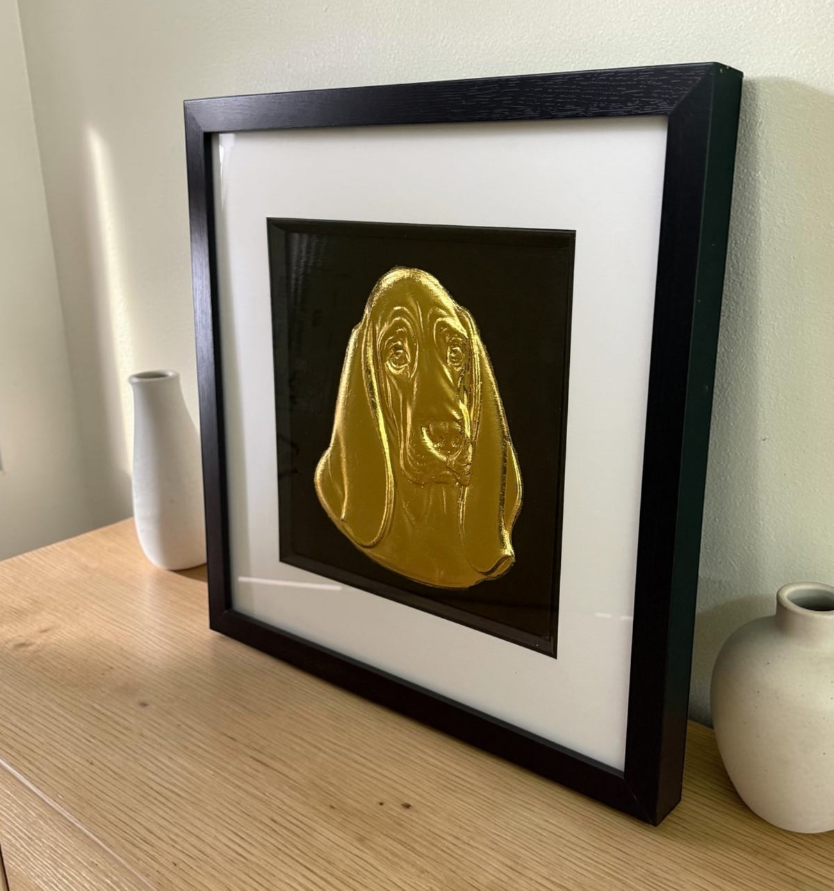Gold Basset Hound framed wall art leaning on an oak sideboard between two white vases, angled to show the raised relief of the ears through the glass