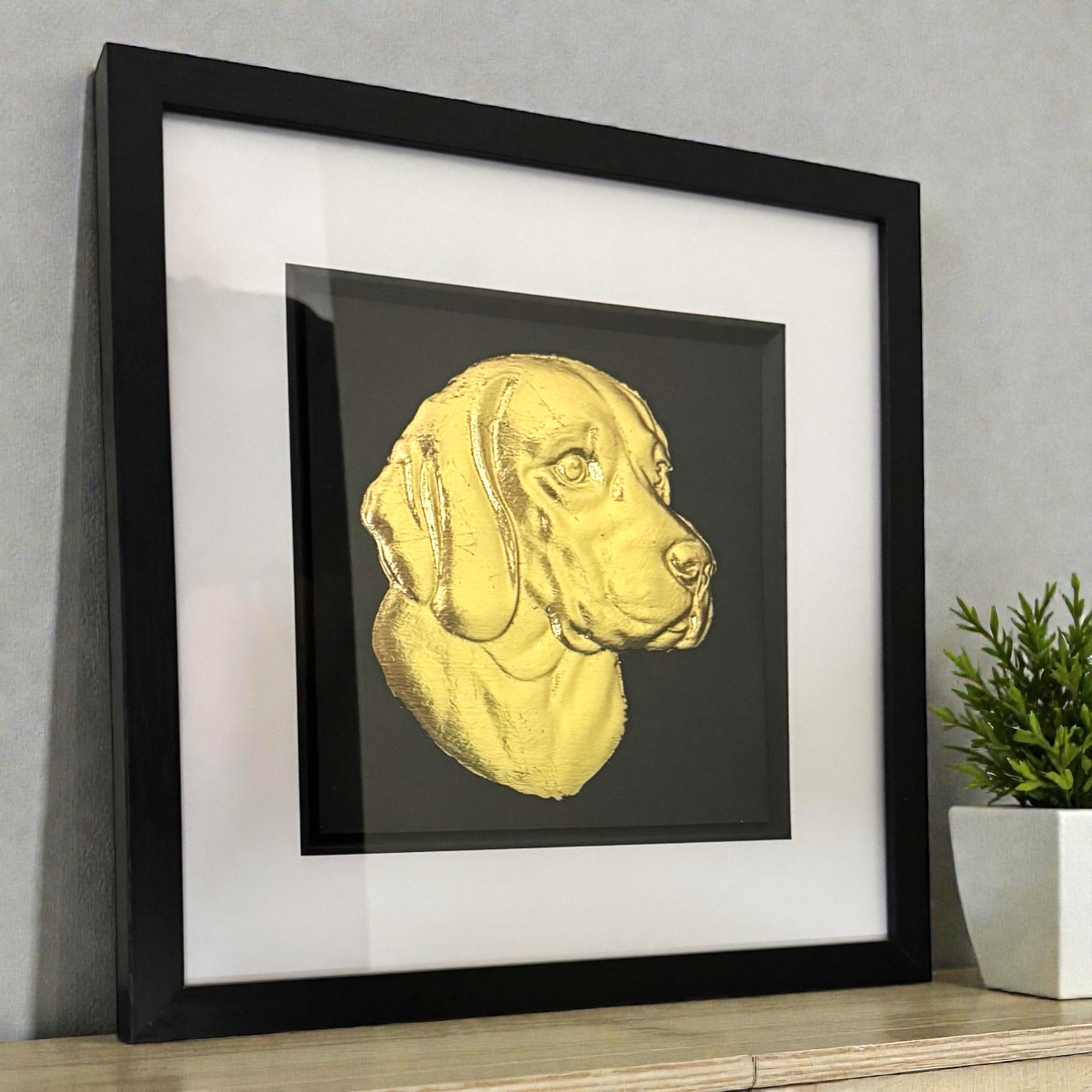 Gold beagle wall art displayed in a black frame with white mount, shown angled on a wooden surface against a grey wall with a small plant for scale