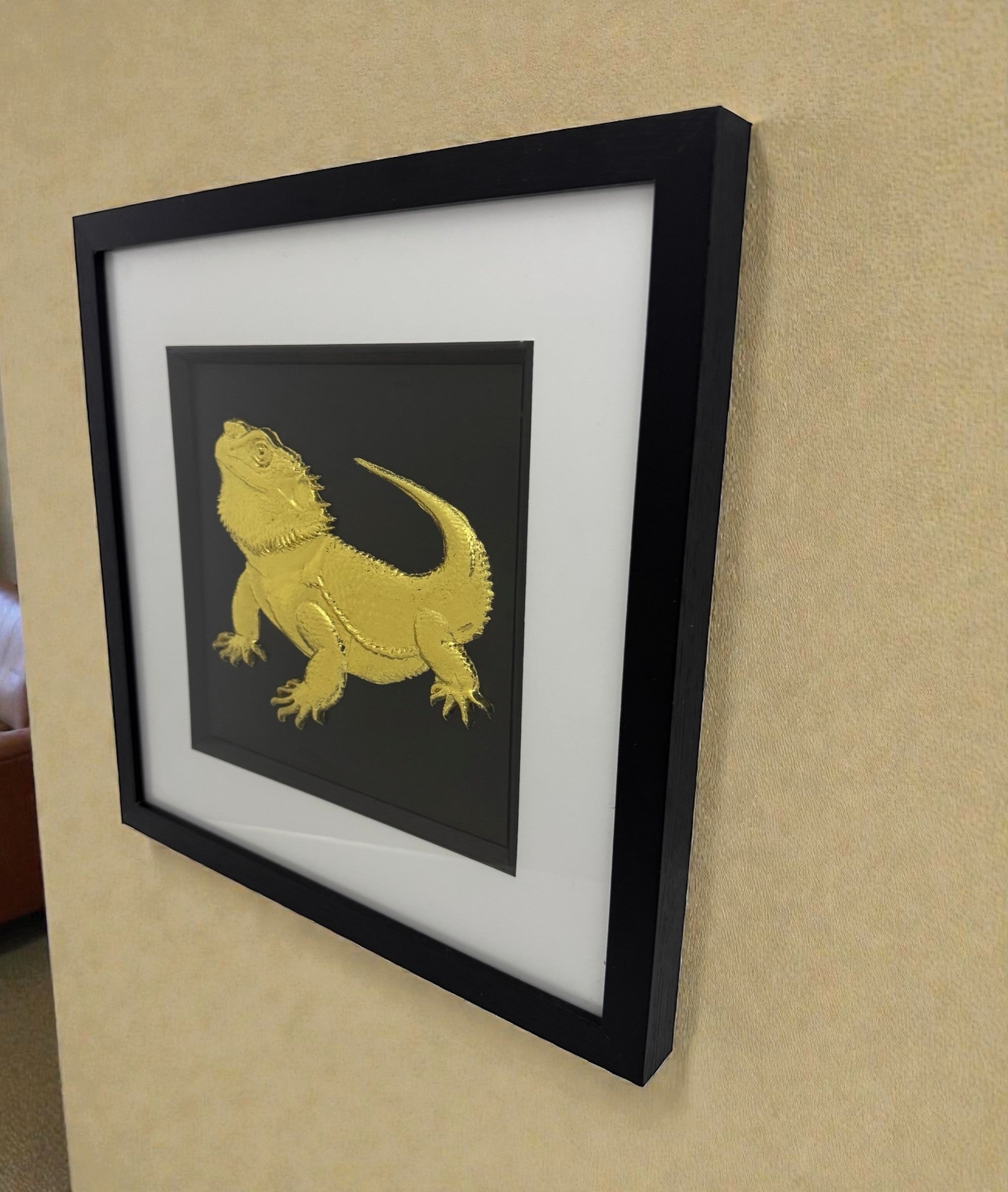 Side view of framed bearded dragon 3D wall art in gold leaf, in a black frame with white mount, hung on a beige wall.