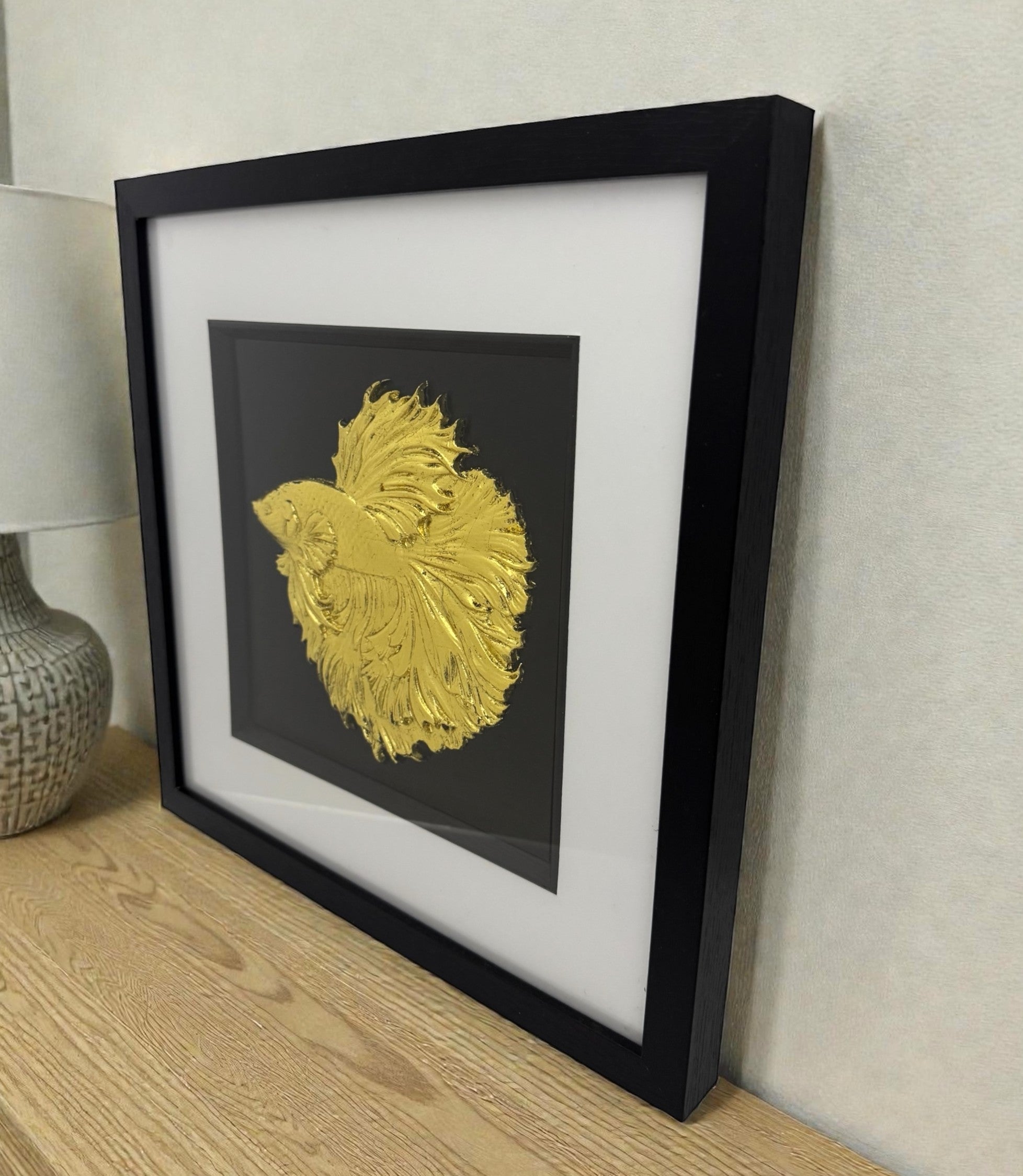 A gold leaf Betta Fish framed art piece displayed on a wooden desk, showing the deep black box frame and the 3D relief texture.