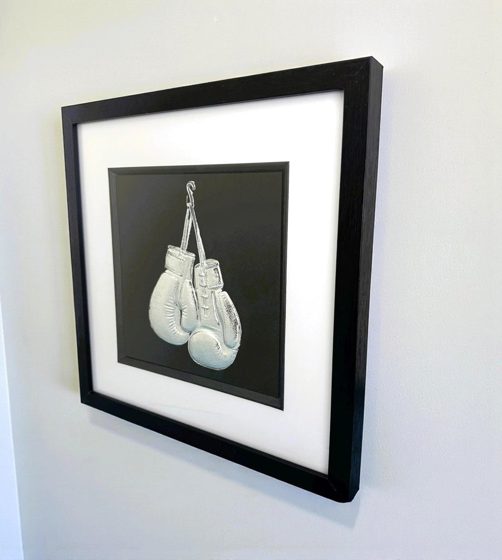 Side view of framed boxing gloves 3D wall art in silver leaf, in a black frame with white mount, hung on a white wall.