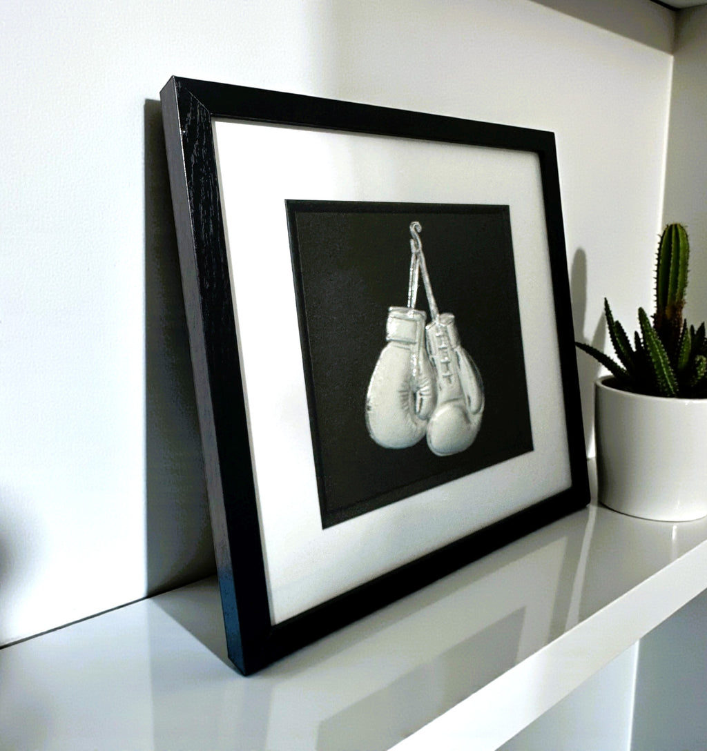 Framed boxing gloves 3D wall art in silver leaf, in a black frame with white mount, styled on a white shelf with a cactus plant.