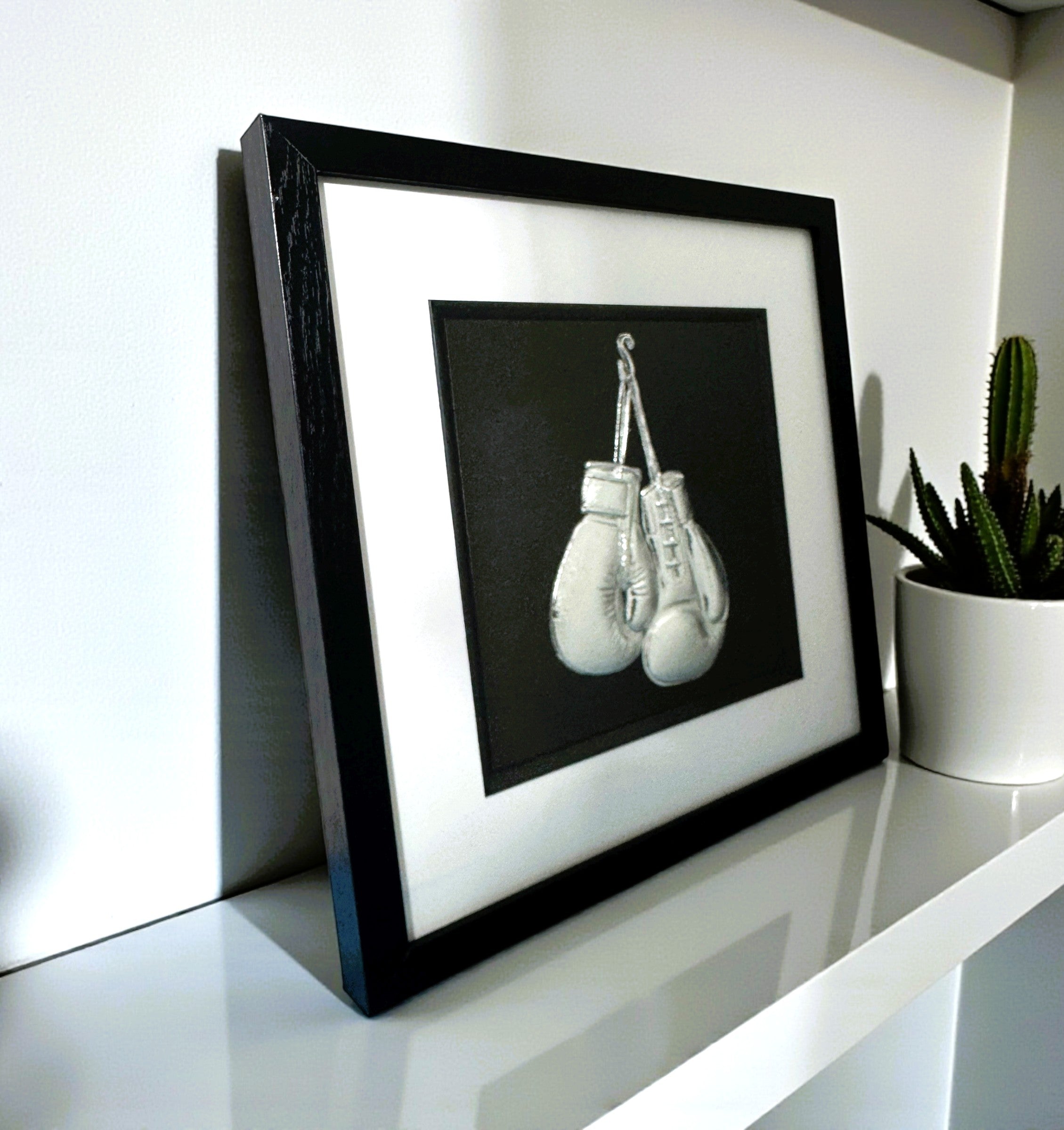 Framed boxing gloves 3D wall art in silver leaf, in a black frame with white mount, styled on a white shelf with a cactus plant.