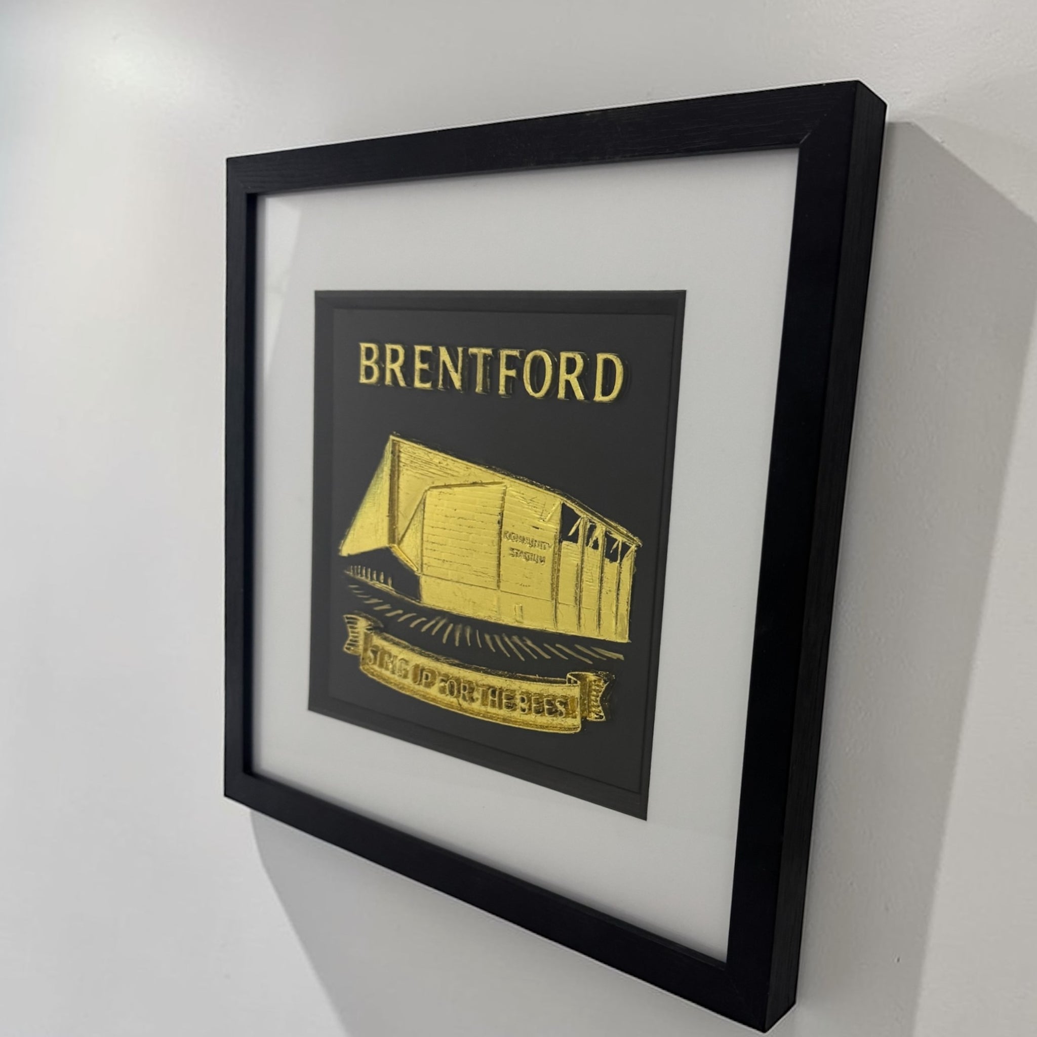 Gold Brentford stadium 3D relief framed artwork on wall, angled side view showing frame depth and glass front