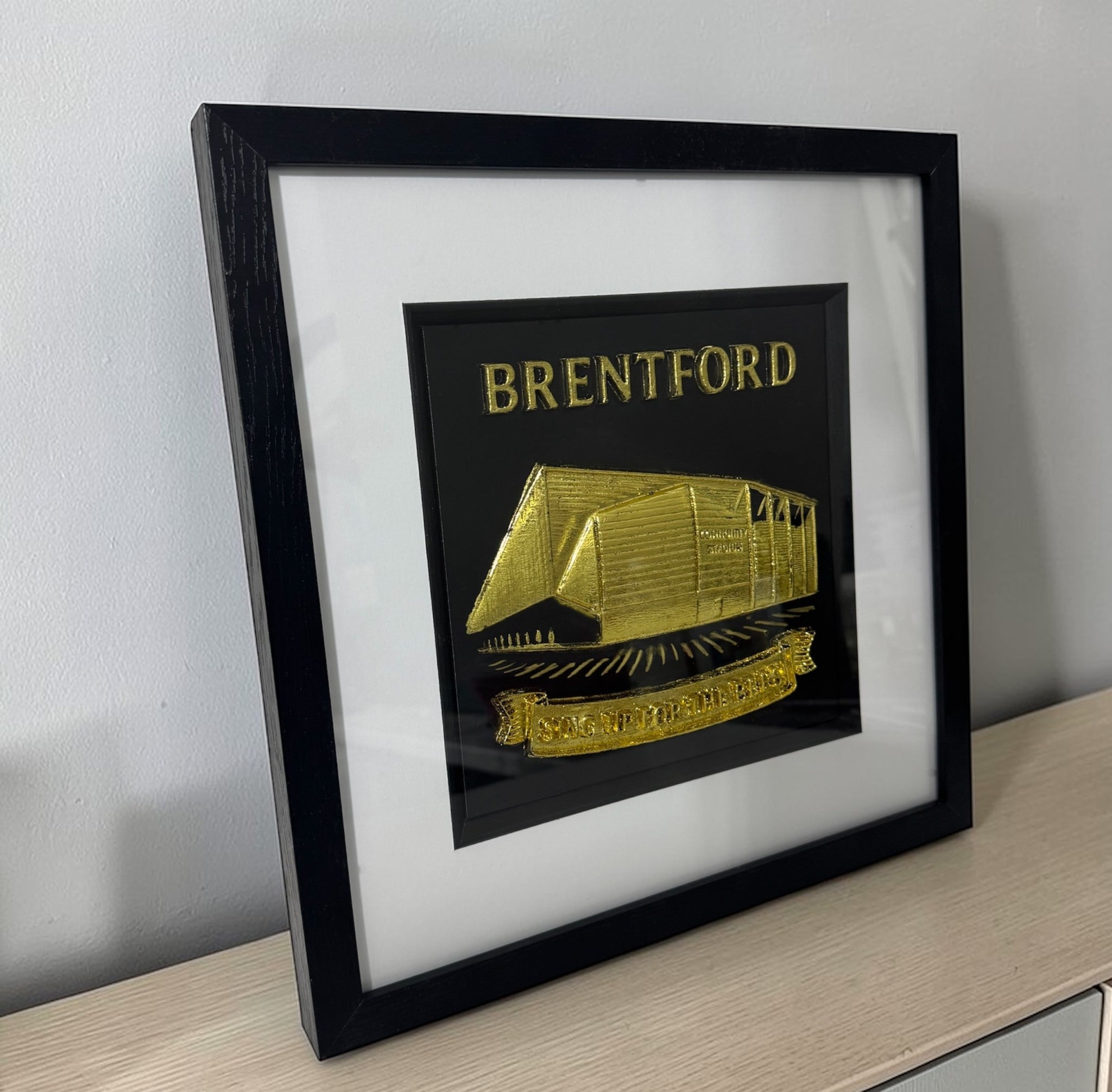 Gold Brentford stadium 3D relief framed artwork on shelf, angled side view showing frame depth and glass front