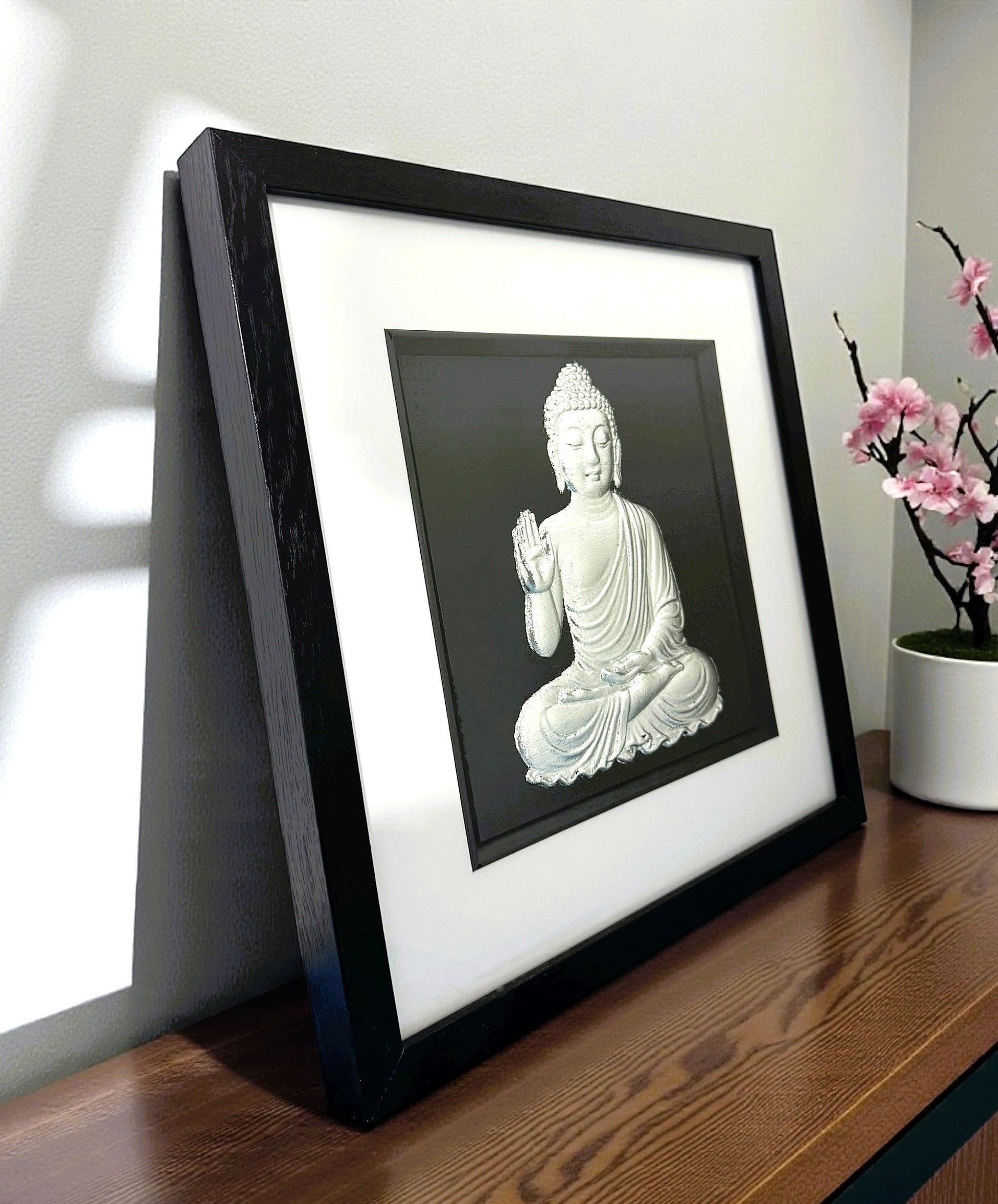 Angled desk view of silver leaf Buddha wall art framed in black with white mount