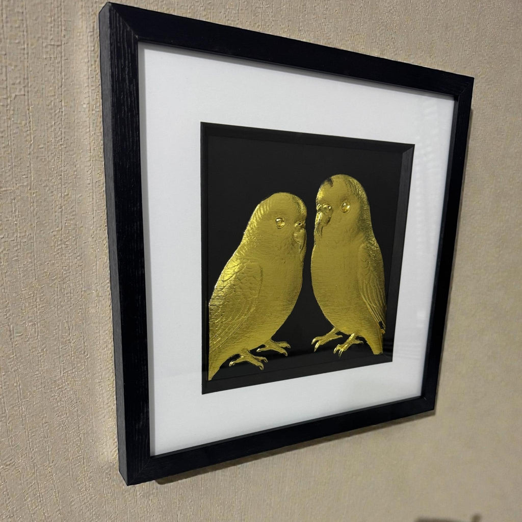 Angled wall view of gold leaf budgie relief artwork in black box frame with white mount on a textured neutral wall