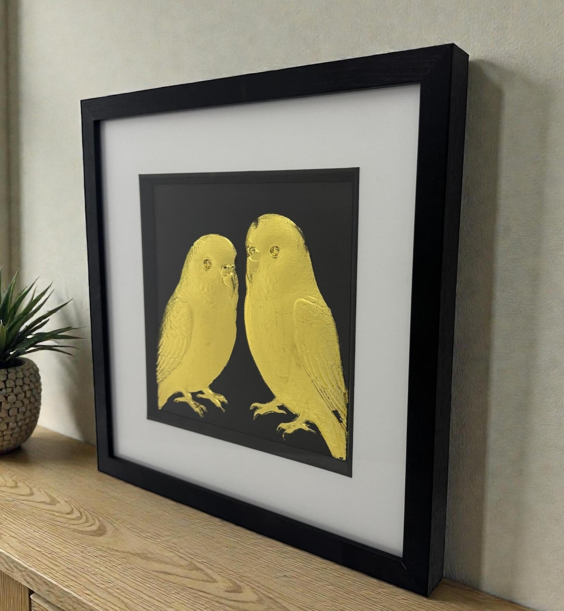 Framed budgie relief artwork in gold leaf, black box frame with white mount, styled on a wooden sideboard against a neutral wall