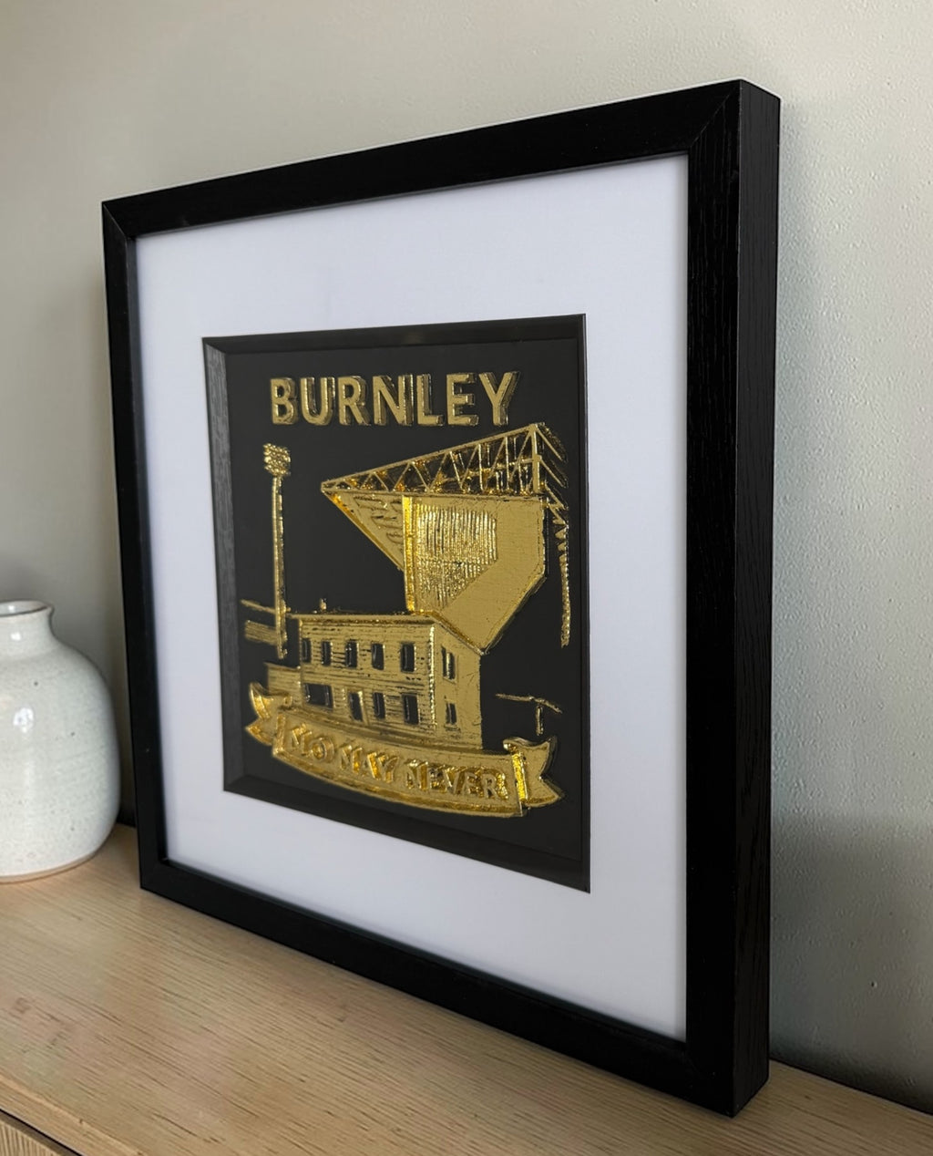 Angled lifestyle image of a gold Burnley football club relief in a black box frame on a wooden sideboard with glass front visible.