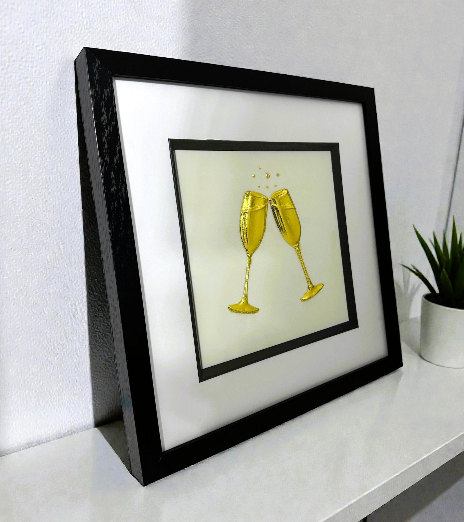 Angled desk view of gold leaf champagne glasses wall art on a light ivory background in a black wooden frame