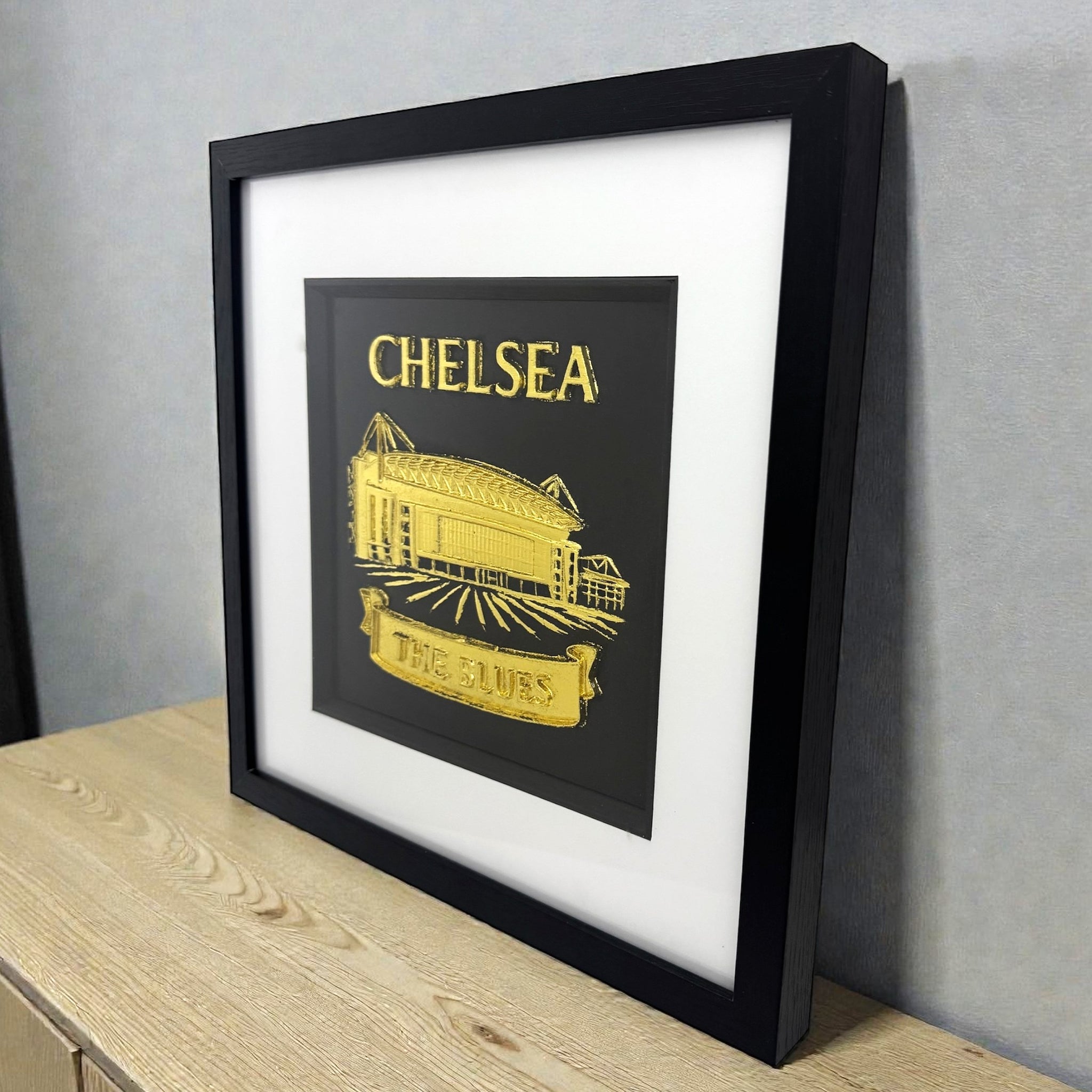 Chelsea stadium wall art in gold leaf shown styled on a desk in a black frame with white mount