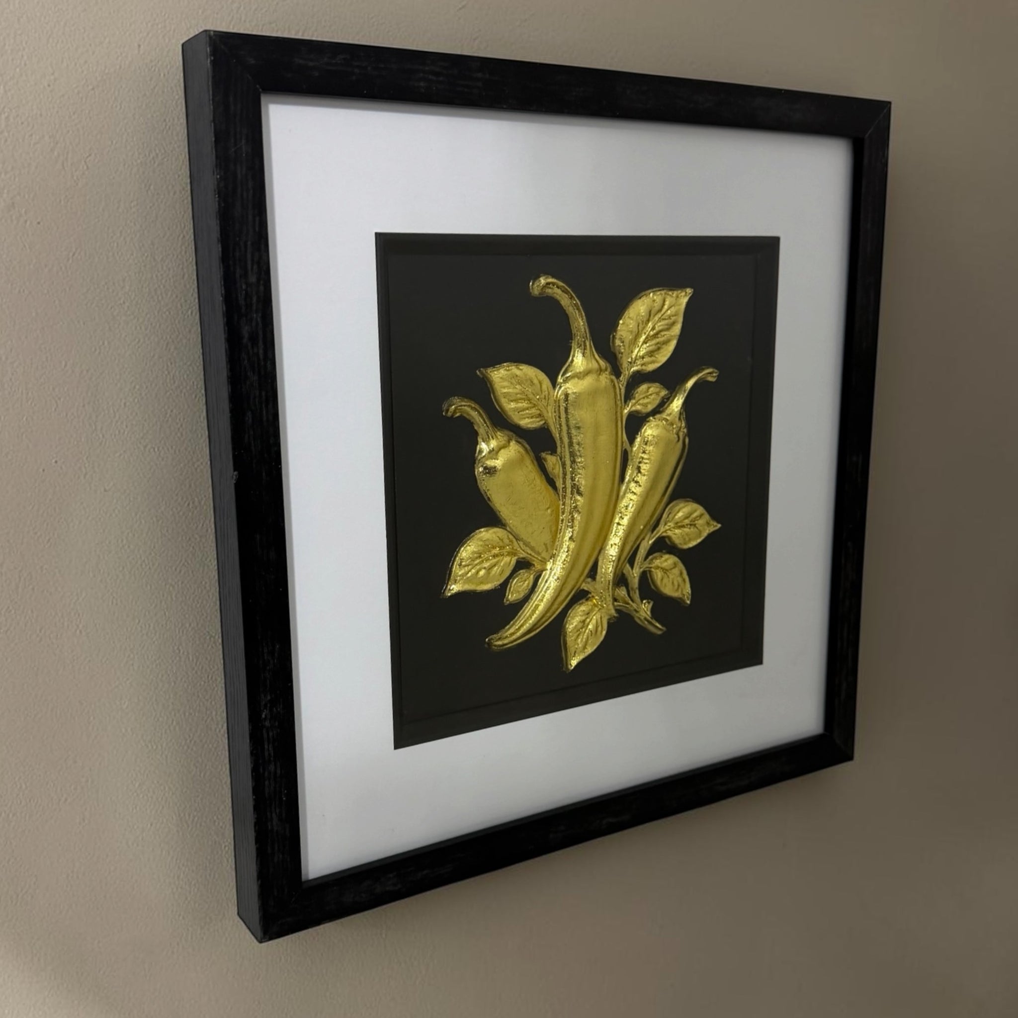 Angled wall view of gold chilli pepper raised relief artwork showing black box frame depth.