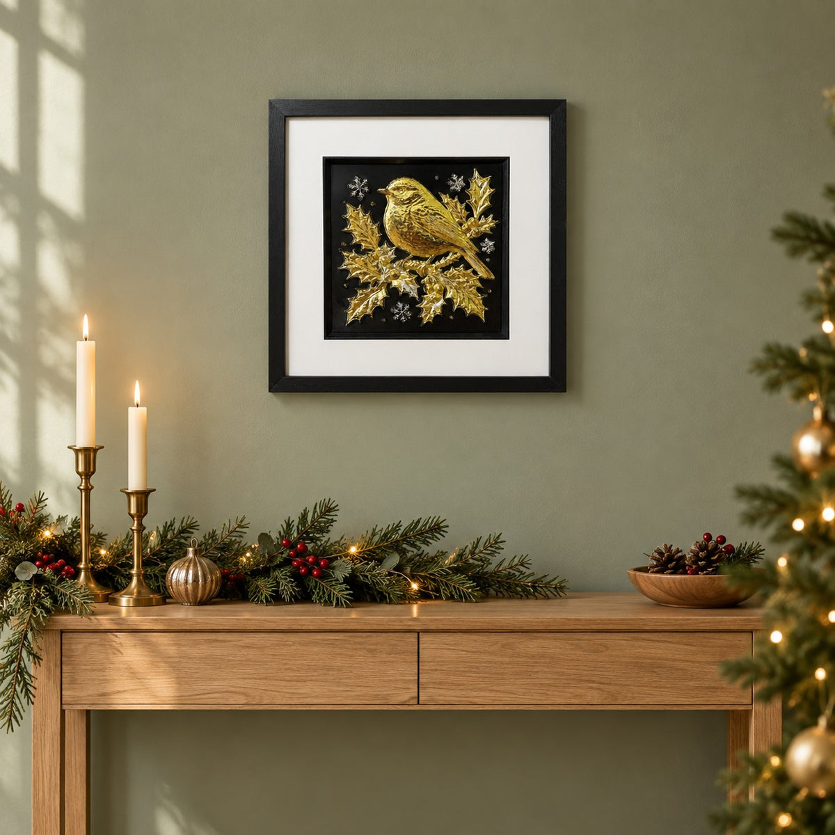 Gold Christmas robin 3D wall art in a black square frame hung on a sage green wall above an oak console dressed with candles, a fir garland and a Christmas tree