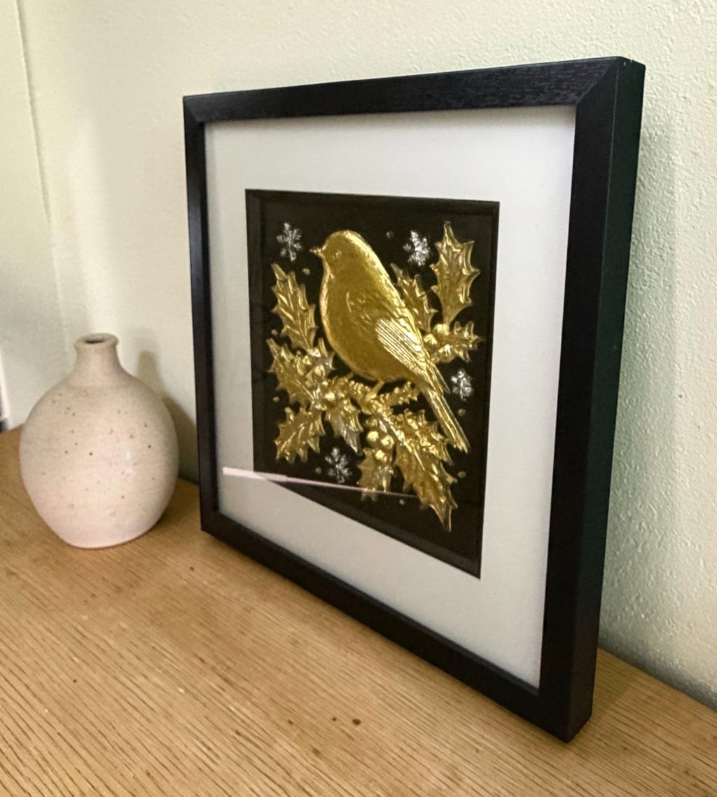 Gold Christmas robin framed wall art with silver snowflakes leaning on an oak sideboard beside a speckled cream vase, angled to show the raised relief through the glass