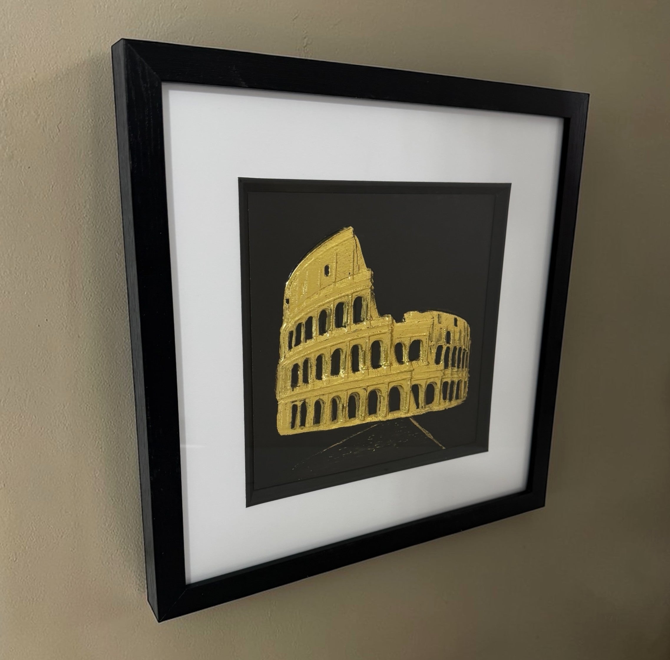 Gold Colosseum raised relief framed in black, shown from an angled side view on a neutral wall.