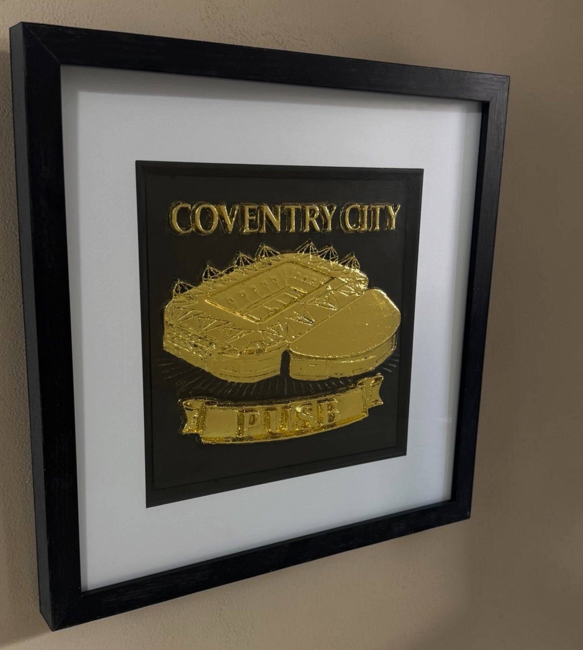 Gold Coventry City stadium framed artwork shown at an angle on a wall in a black square box frame